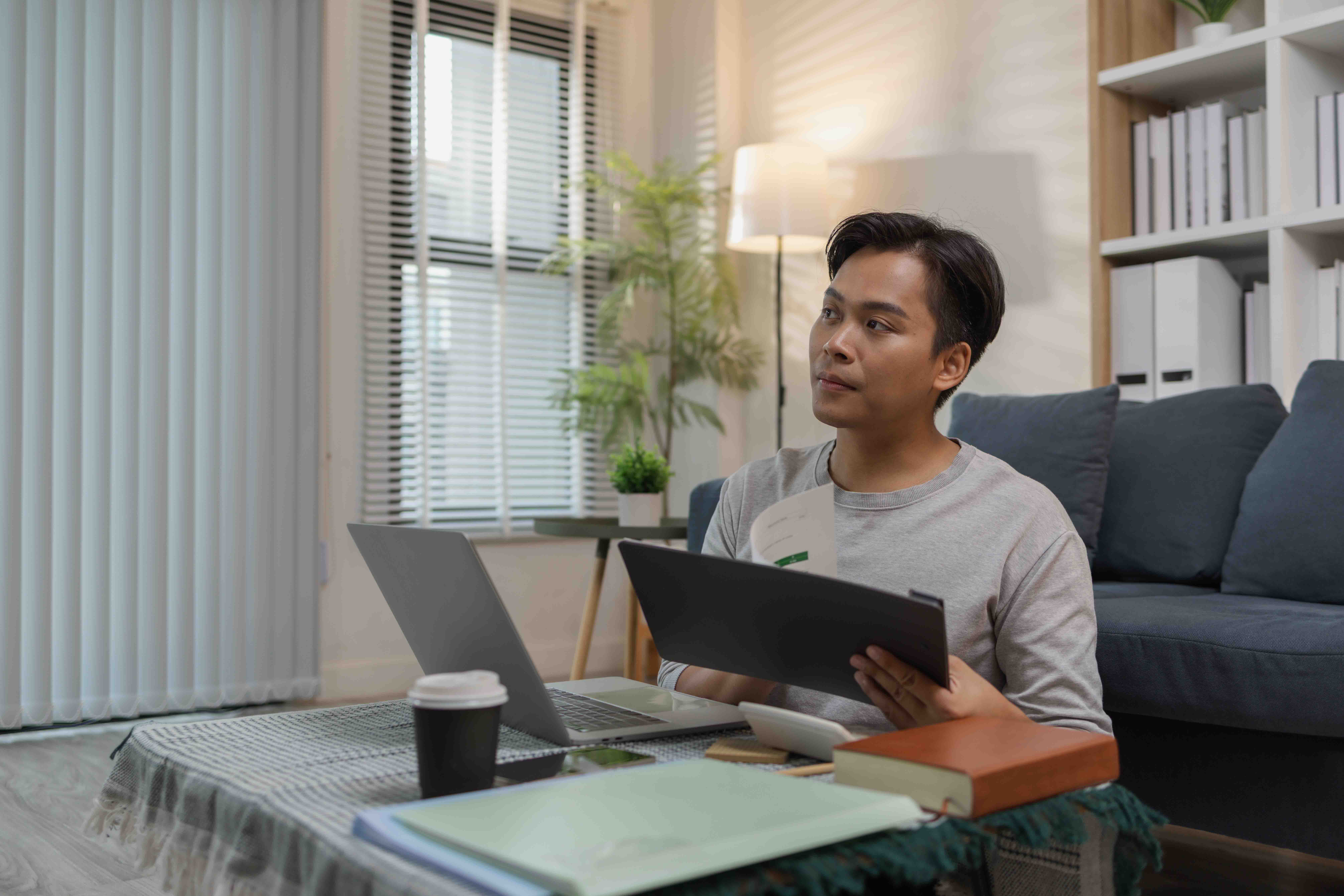 A young man is sitting on the floor at home, checking documents while working with his laptop. He appears focused and engaged, managing his tasks in a relaxed modern living space.