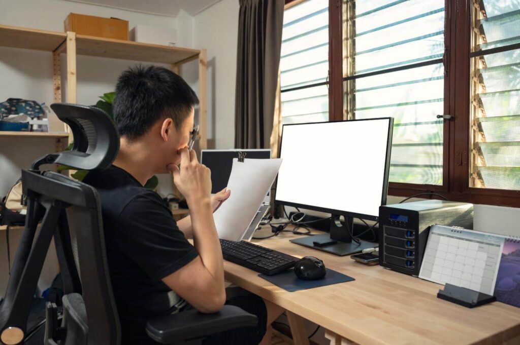 freelance-young-asian-man-working-and-examining-paperwork-and-monitor-display-at-wooden-table