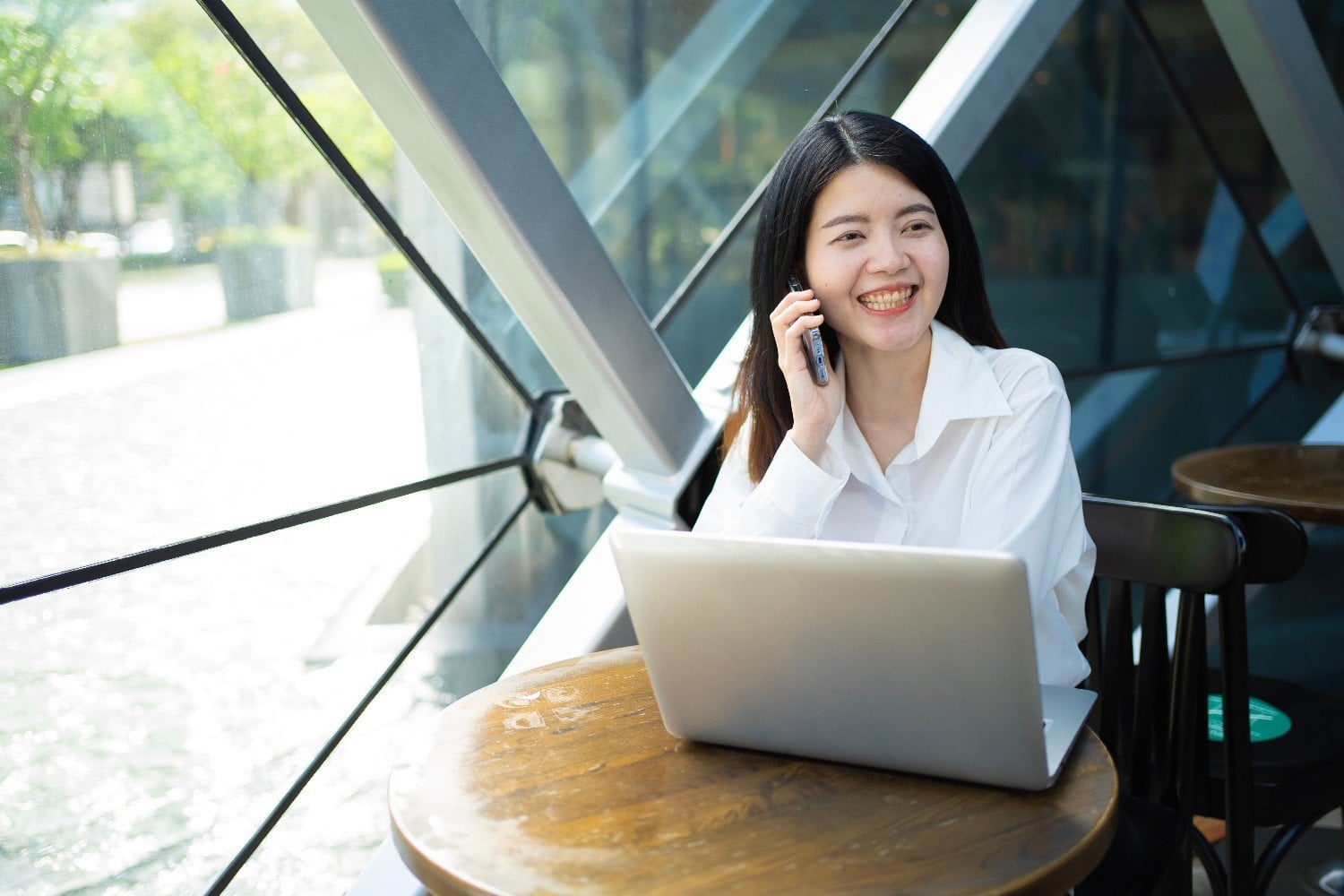 Smiling virtual assistant taking a client phone call at a laptop by a cafe window