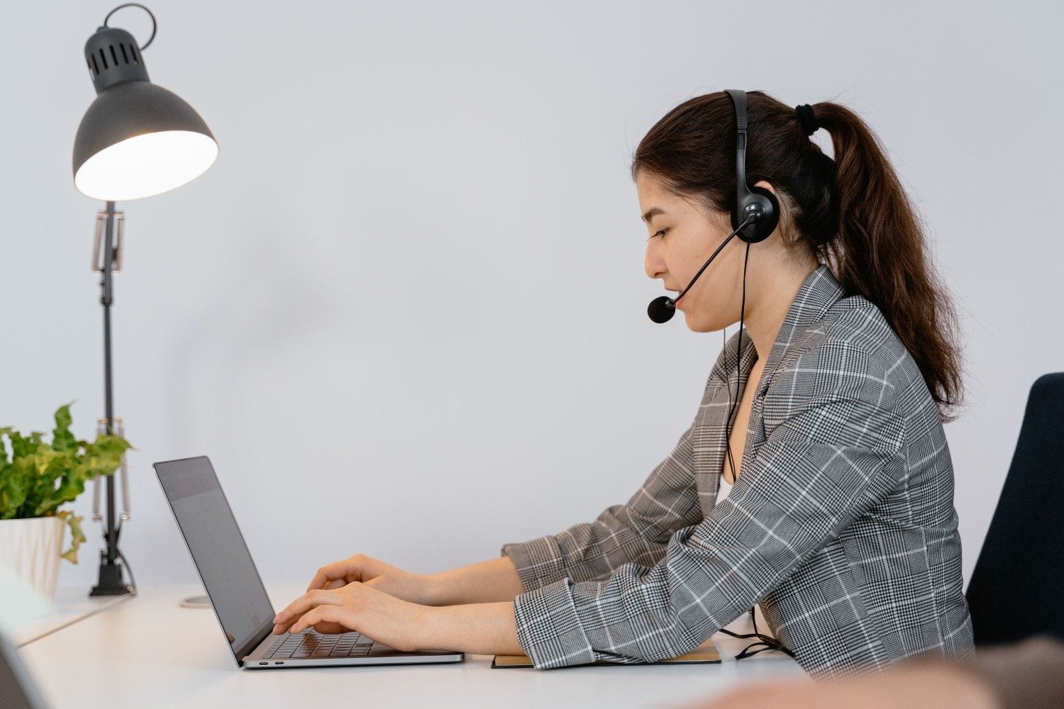 Virtual assistant in a headset typing on a laptop at a desk lamp-lit workstation